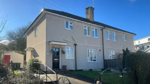 A property on Cobhorn Drive in Bristol. The flat is beige in colour with several windows on the front. The front door is black and to the side. There are hanging baskets either side of the door.