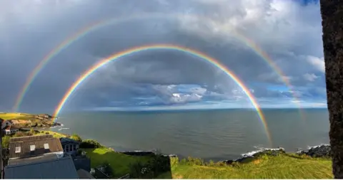 Lorraine Sharkey A striking coastal landscape photograph featuring a double rainbow over the sea. Several buildings with grey and brown roofs are visible in the lower-left corner while the double rainbow arcs against dark clouds.