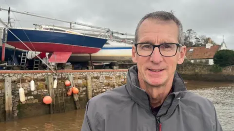 BBC Commodore Ian Garcia is wearing black-framed glasses and a grey waterproof jacket with boats in Topsham Sailing Club boatyard visible behind him.