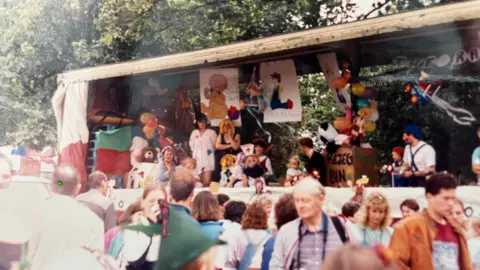David Edwards A carnival with lots of people in colourful outfits on the back of lorry trailer.