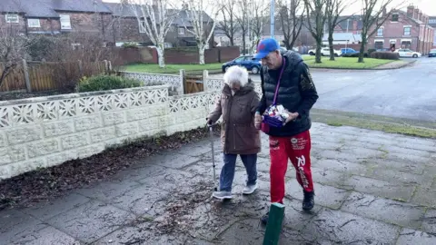 June Horvath is helped to walk by Paul Wilson. She has a brown coat and blue trousers, while he has a blue cap and top and red trousers.