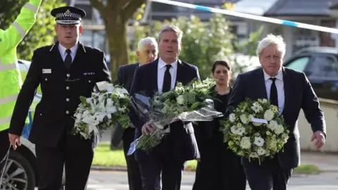 Downing Street/EPA Ben-Julian Harrington, Sir Keir Starmer and Boris Johnson, all smartly dressed in suits, carrying large bouquets of flowers. They are walking past a police cordon on a residential street.