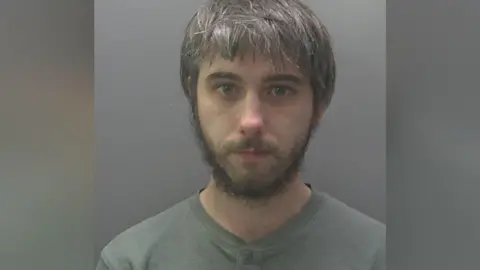 Joshua's custody image. He has black hair going grey and a black beard. He is wearing a grey t-shirt.