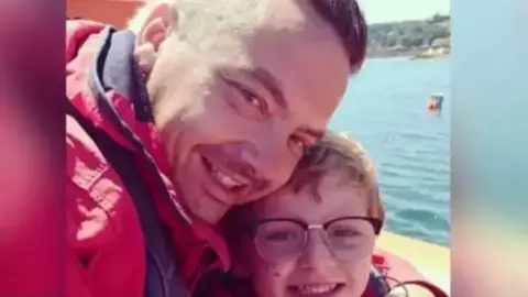 Family handout A man and son holding each other close and smiling to the camera in a selfie style, with the sea behind them. The young boy is wearing glasses and the father a large red coat.