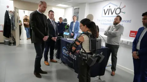 PA Media Prince William looks at a woman covered in an exoskeleton fitted with large battery packs. The prince wears a black suit and black sweater, A number of other people are looking on. Writing on the wall says VIVO. The UWE branding is also visible. 
