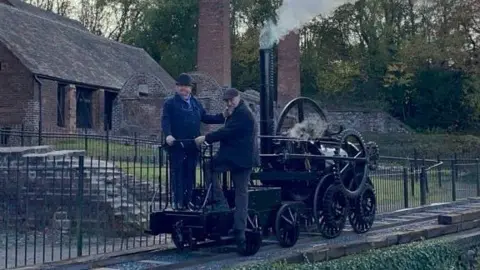 Two men are standing on a small black steam engine which is on a small railway line. It has a long, tall chimney with steam coming out of it, and large wheels on the side of it.