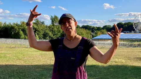 A woman standing in an empty field at a festival. She is wearing a dark purple top and a cap. She is holding both of her arms up and using her fingers sign the "rock on" symbol, with her thumb, index finger and little finger up. There is a white big wheel in the background and the sky is blue.