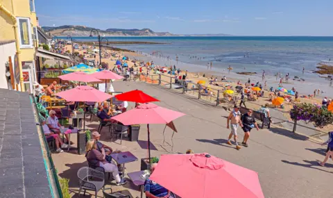 Love Lyme Regis View looking down on cafe tables and red umbrellas outside a cafe on the promenade at Lyme Regis. It's a sunny summer's day and people are sitting at the tables, walking along the promenade and the beach and shore is packed with people.