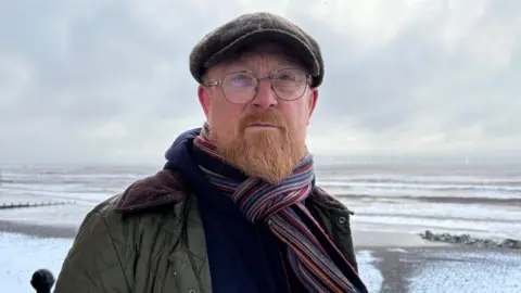 Kevin Shoesmith/BBC A man with a ginger beard and wearing glasses and a flat cap looks at the camera. Behind him is a snow-covered beach.