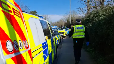 A police officer walking on a pavement in a high-visibility vest. There are multiple police cars parked on the side of the road.