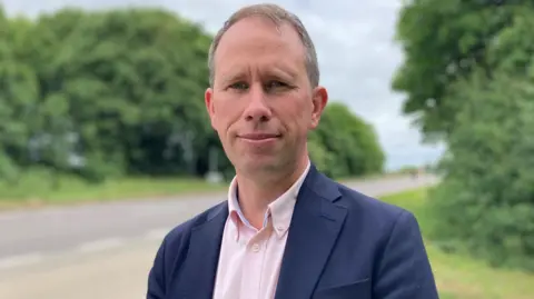 Matthew Braber has short brown hair and is standing next to a road whilst wearing a blue blazer over a pink shirt.