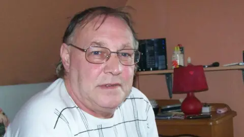 Sussex Police A man wearing a white t-shirt with black lines on it, also wearing glasses. He is sat in a room with brown walls, a wooden table and a red lamp in it.