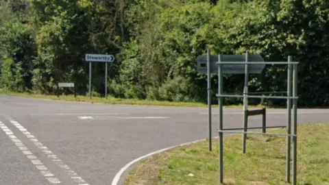 Google A road T-junction. A white sign printed with the word STEWARTBY points to the right of the frame, in the direction of a grey concrete road.