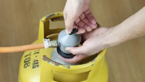 Two hands are operating a silver valve on a yellow gas canister, which is on a wooden floor. An orange hose is attached to the canister.