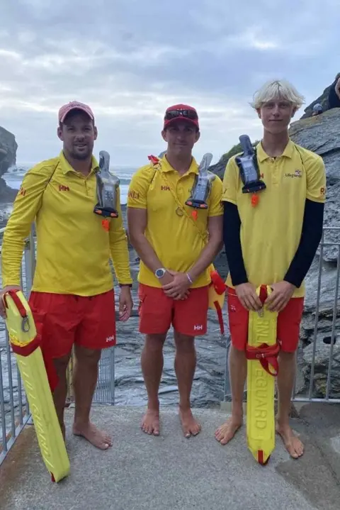 RNLI RNLI lifeguards Andy Boxall, Sam Leader and Ollie Keast stood in a line wearing their yellow and red uniform while holding safet equipment.