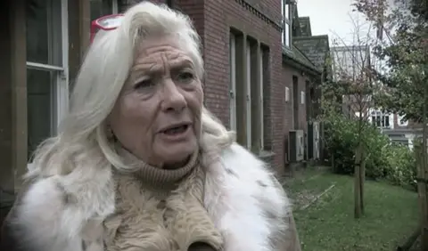 Dorothy Hayes, who has long white/blonde hair and is wearing a beige coat with a white fur collar. Underneath she had on a high-necked beige jumper. She is pictured from the chest up, if looking to the left of frame and is standing beside some buildings by a patch of grass with trees in it.