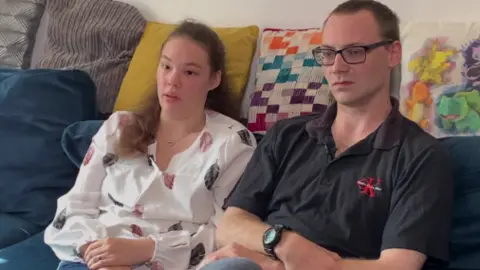 Couple affected by high rental costs who were forced to move to a smaller house sitting on a sofa