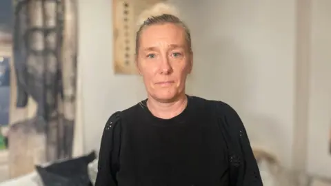 Kelly Lees has short light hair. She is looking directly towards the camera and wears a black jumper