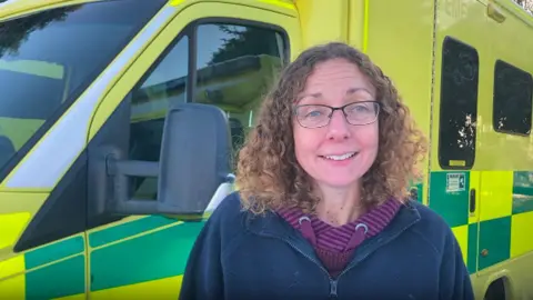 Head and shoulders image of Julie Rowlandson standing in front of an ambulance. She is smiling, has shoulder length, brown, curly hair and is wearing glasses and a navy fleece