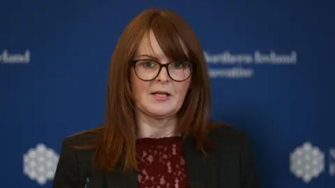 PA Media A ginger haired woman stands in front of a blue wall. She wears a grey suit with a red top. She wears brown glasses. 