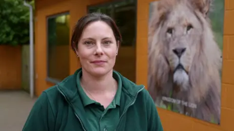 Shaun Whitmore/BBC Frankie is standing outside in front of a lion poster and smiling. She is wearing green zookeeper clothing.