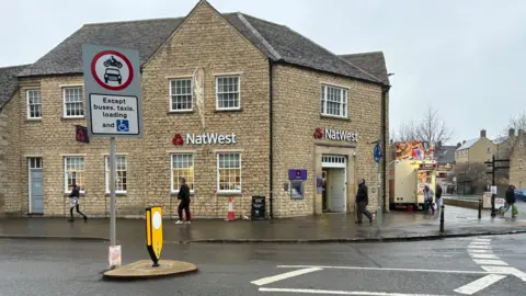A large traffic sign stands at the start of the high street. On it is a car and motorbike symbol in a red circle stating those vehicles can't enter. Below the picture is writing which says 'Except buses, taxis, loading and the disabled symbol.' Behind the sign is the bank Natwest where people can be seen walking in front of it.