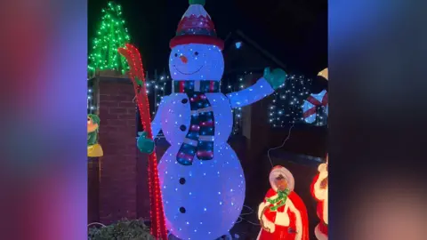 Paul Molyneux A large snowman figure shines brightly with white lights, wearing a red hat and striped scarf. The snowman holds a red ski and is surrounded by other festive decorations. Green and blue lights add color to the scene.