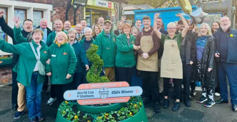 Members of staff and volunteers celebrate the win, standing outside the station entrance and in front of a floral display in a green planter. A sign saying 'World Cup of Stations 2024 Winner' is on top of the planter. Everyone is smiling. Some people are raising their arms in the air.