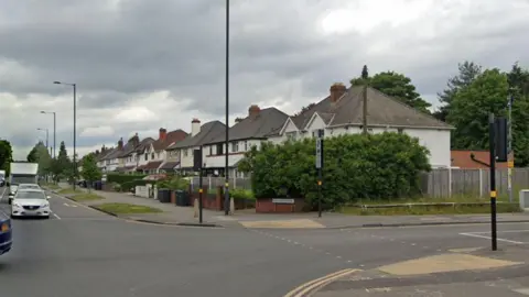 A road junction with a row of white houses running down one side. and a number of vehicles on the road waiting to get through the crossroads.