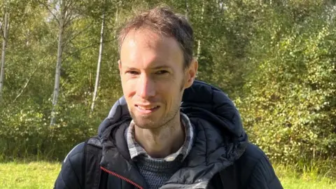 Ollie Conopo/BBC Man with short brown hair wearing a blue jacket stands in a field