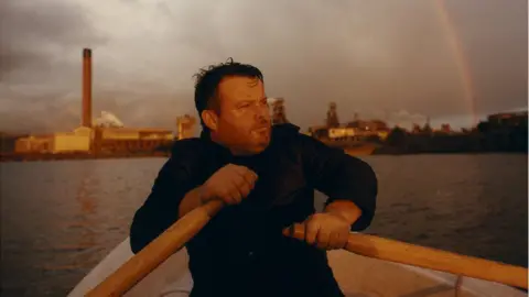 Roo Lewis Michael Locke from Dirty Sanchez in a rowing boat in Port Talbot in front of the steelworks