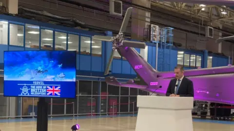 A man on a podium speaking in front of a purple helicopter