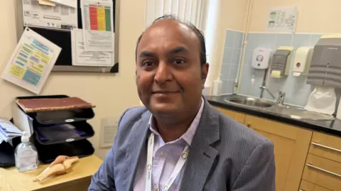 A man in a GP surgery room wearing a grey suit jacket