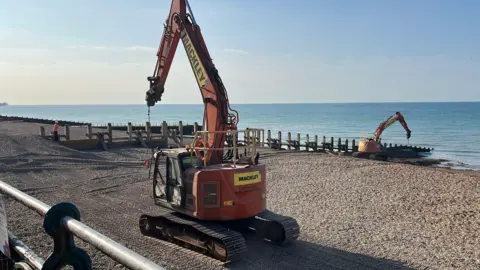 The image shows two building contraptions on an empty beach, with the sea visible in the right-hand-side of the image.