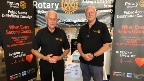 Two men stood by boards campaigning for defibrillators. They are both elderly, have white hair and are wearing black polo shirts with yellow Rotary Club logos. 