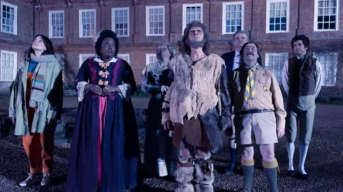 A still from the sitcom Ghosts which sees the cast standing outside in the dark and looking up at the sky. The estate house can be seen in the background.
