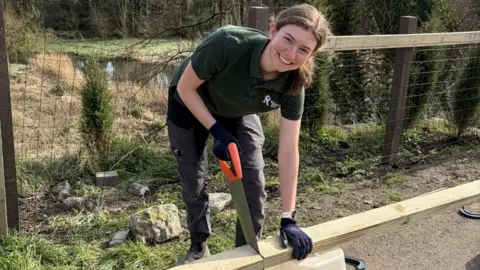 Rosie Ashworth A young woman with brown hair tied back in a ponytail saws into a plank of wood. She wears a green polo shirt, black cargo trousers, working boots and gloves.