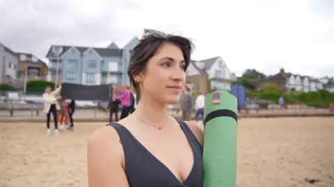 A woman with a black vest and on holding a green yoga mat, she is standing on a sandy beach with other people and houses in the blurred background.