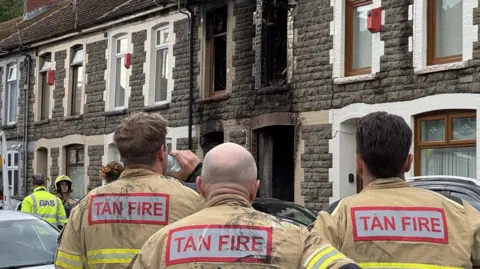 BBC A stone terraced house with fire damage around the windows and doors. Three fire officers look at the house with their backs to the camera.