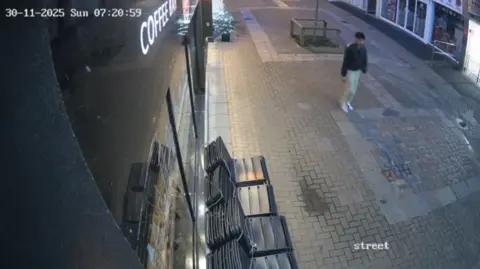 Jersey Police CCTV image of a pedestrianised street outside a coffee shop in the early morning. Abdelkarim Arfaoui is wearing a dark jacket, light-coloured trousers and trainers, he is walking along the paved street. Stacked outdoor chairs are lined up against the coffee shop window in the foreground. Shopfronts and street furniture are visible in the background. A timestamp in the top-left corner reads “30-11-2025 Sun 07:20:59”.