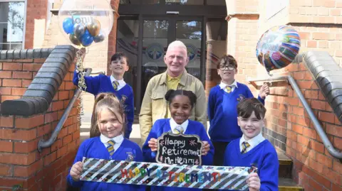 A man with short white hair, wearing a yellow polo shirt underneath a light brown jacket stands on the steps in front of a school building. With him are five children, wearing white shirts and yellow-and-blue-striped school ties underneath school sweaters. Two are holding a banner reading "HAPPY RETIREMENT", another is holding a sign reading "Happy Retirement Mr Henry", and two are holding balloons. All are smiling at the camera.