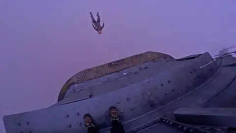 A still image taken from a video of a base jumper at the top of the Humber Bridge. It has been taken from a first-person perspective and shows a man jumping from the structure into a misty sky. The photographer is standing on a grey metal ledge and their feet are visible.