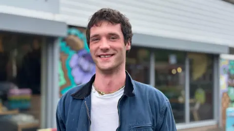 Gemma Dillon/BBC Image shows a man wearing a white t shirt with a blue jacket.
He is standing on the street in Meanwood with a coffee shop behind him.
