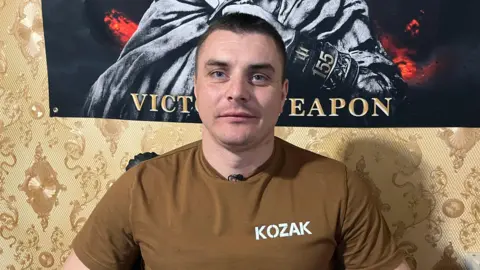 BBC A man in a khaki t-shirt with the word 'Kozak' written on it stares directly at the camera