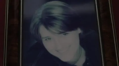 A framed photo of a dark-haired woman's face. She is smiling and is wearing a necklace and a dark-coloured top.