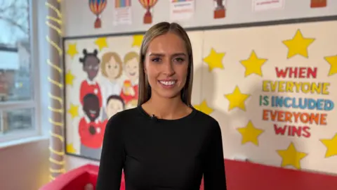 Rachel Donnelly wearing a black top stands in a brightly decorated classroom area. Behind them is a wall display with yellow stars and the slogan “WHEN EVERYONE IS INCLUDED EVERYONE WINS.” To the left are colourful drawings of children in school uniforms, and to the far left is a large window with string lights wrapped around the frame. A red soft‑play structure is partially visible at the bottom of the image.