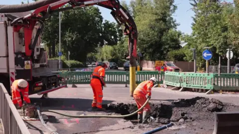 Workers in high-vis use large machinery to dig up a road while behind temporary barriers.