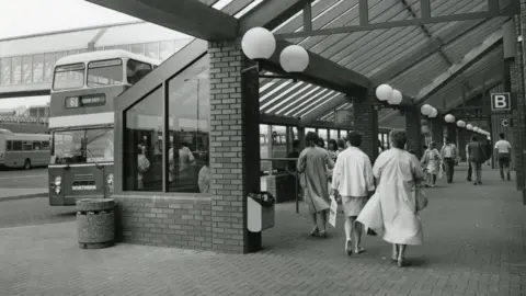 Metrocentre A black and white photograph of the bus station with a double decker bus on the right. On the right women dressed smartly are walking alway from the camera towards the stands. The woman on the right is wearing a trench coat and the woman next to her on the left is wearing a pencil skirt and jacket.