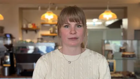 A woman with blonde hair and wearing a knitted jumper. She is standing in a cafe, the counter and some lighting is visible in the background.