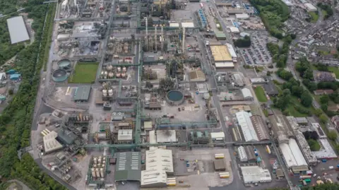 An aerial view of an industrial site with lots of large buildings, surrounded by trees and houses.